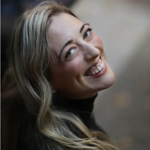 A portrait of Kristel Kubart: she looks back at the camera with a large smile. Her long blonde hair falls down by her right shoulder.