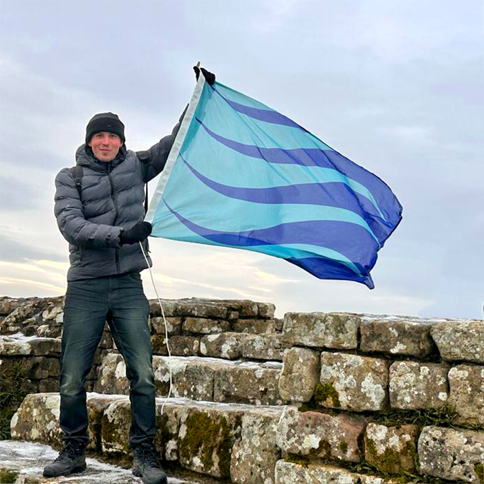 A photo of Ramdeep Roman standing atop Hadrian's Wall, waving this Stuttering Pride Flag.