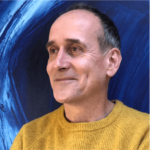 A blue-tinted portrait of Paul Aston: he is wearing a bright yellow jumper. He is looking off into the distance with a smile, his face turned at a three-quarters angle against the camera. Behind him is a painterly blue backdrop.